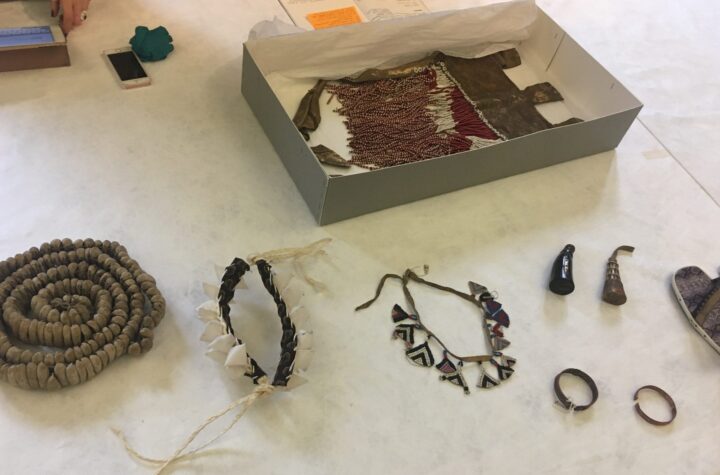objects from Botswana laid out on a table with a white Tyvek cover. One object, a red and white beadwork apron, is shown in a grey conservation box.
