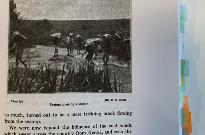 Page in a book which shows a pixelated black and white photograph of several men carrying heavy equipment over a river with the caption 'Porters crossing a stream'.