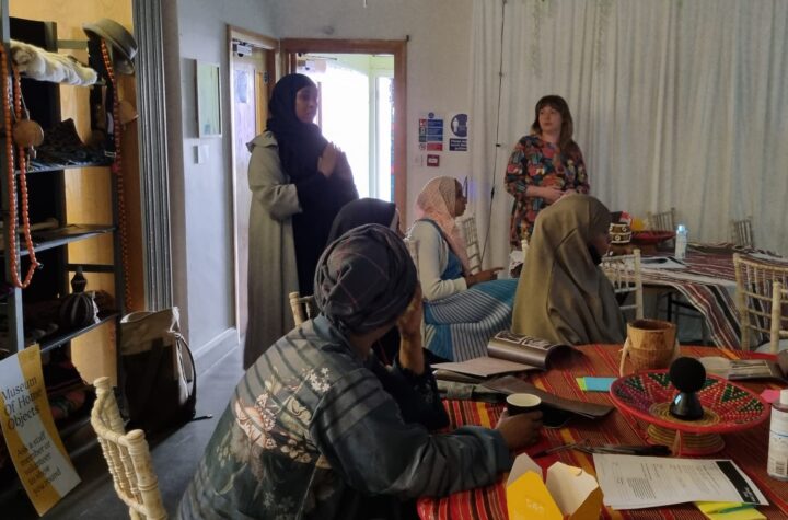 a user research workshop taking place at a community centre. Two women are standing and talking, other women sit listening and discussing. There are shelved with Somali cultural objects in the background.